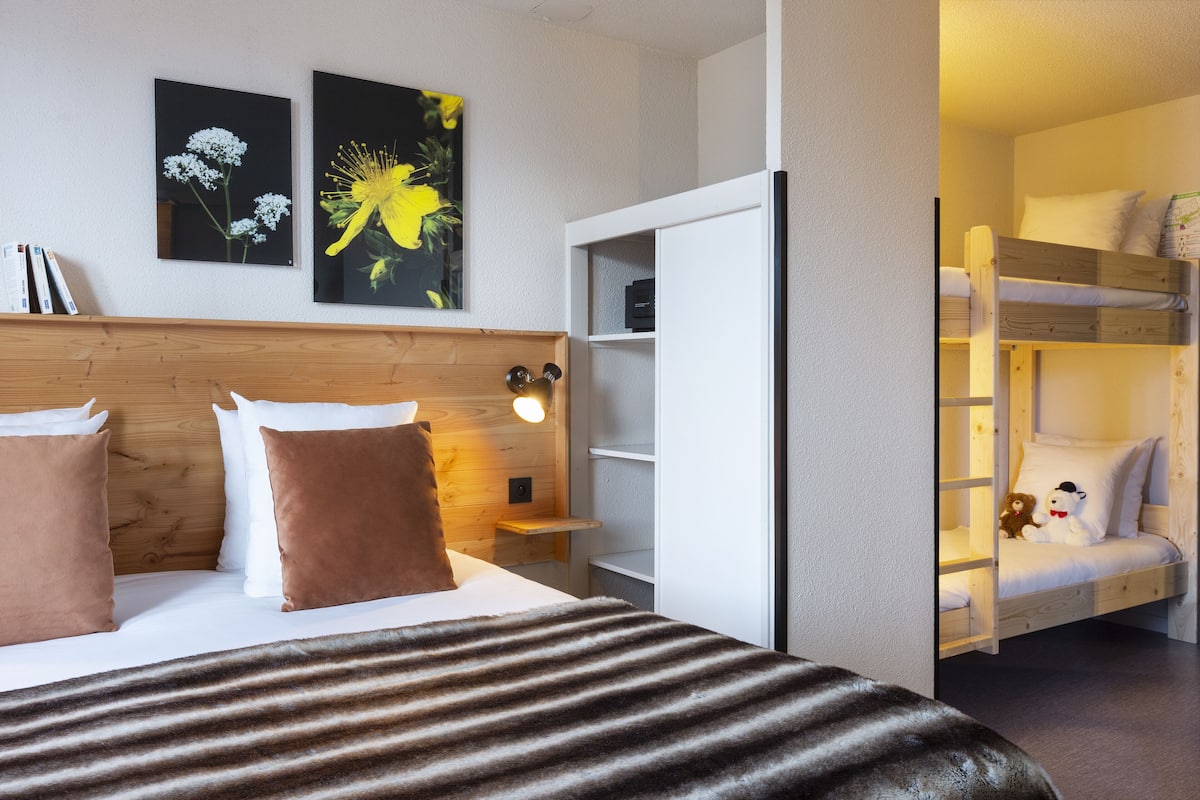 Habitación de hotel con cama, cabecera de madera, mesita de noche con lámpara, estantería y una litera con peluches.