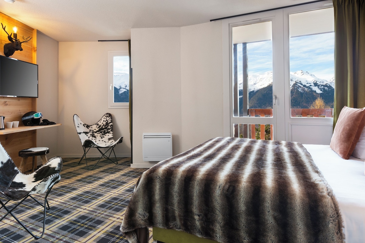 Habitación de hotel con cama, una silla, un escritorio, televisor y ventana con vista a la montaña.