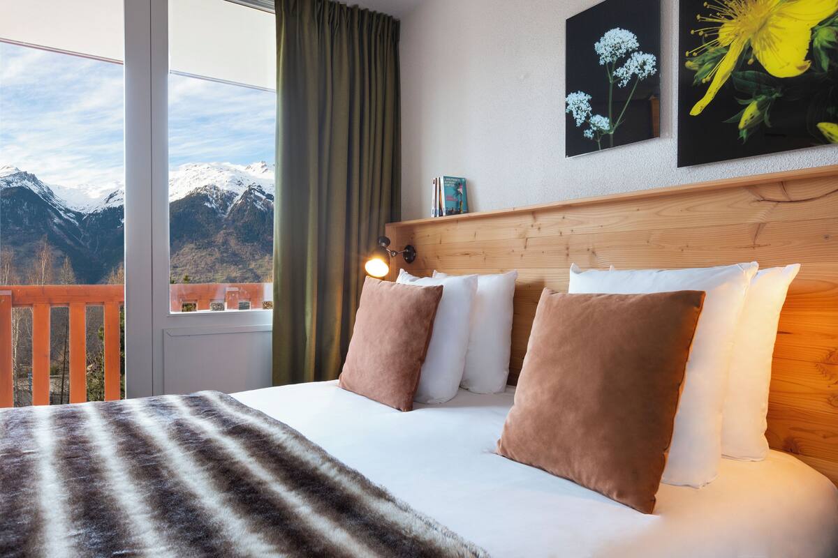 Una cama con vistas a montañas a través de una ventana, cabecera de madera y un cuadro de flores.
