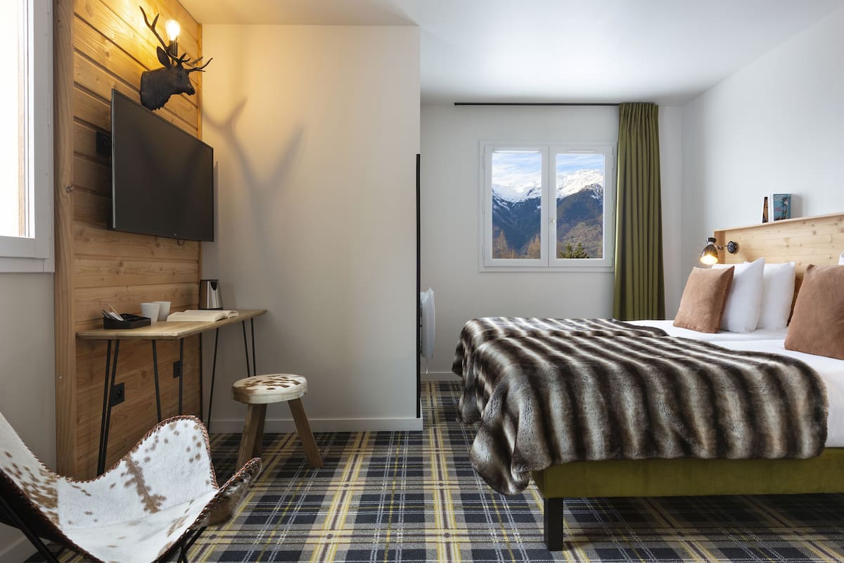 Un dormitorio con cama, televisor montado, escritorio de madera, silla, ventana con vista a la montaña y alfombra de estampado escocés.