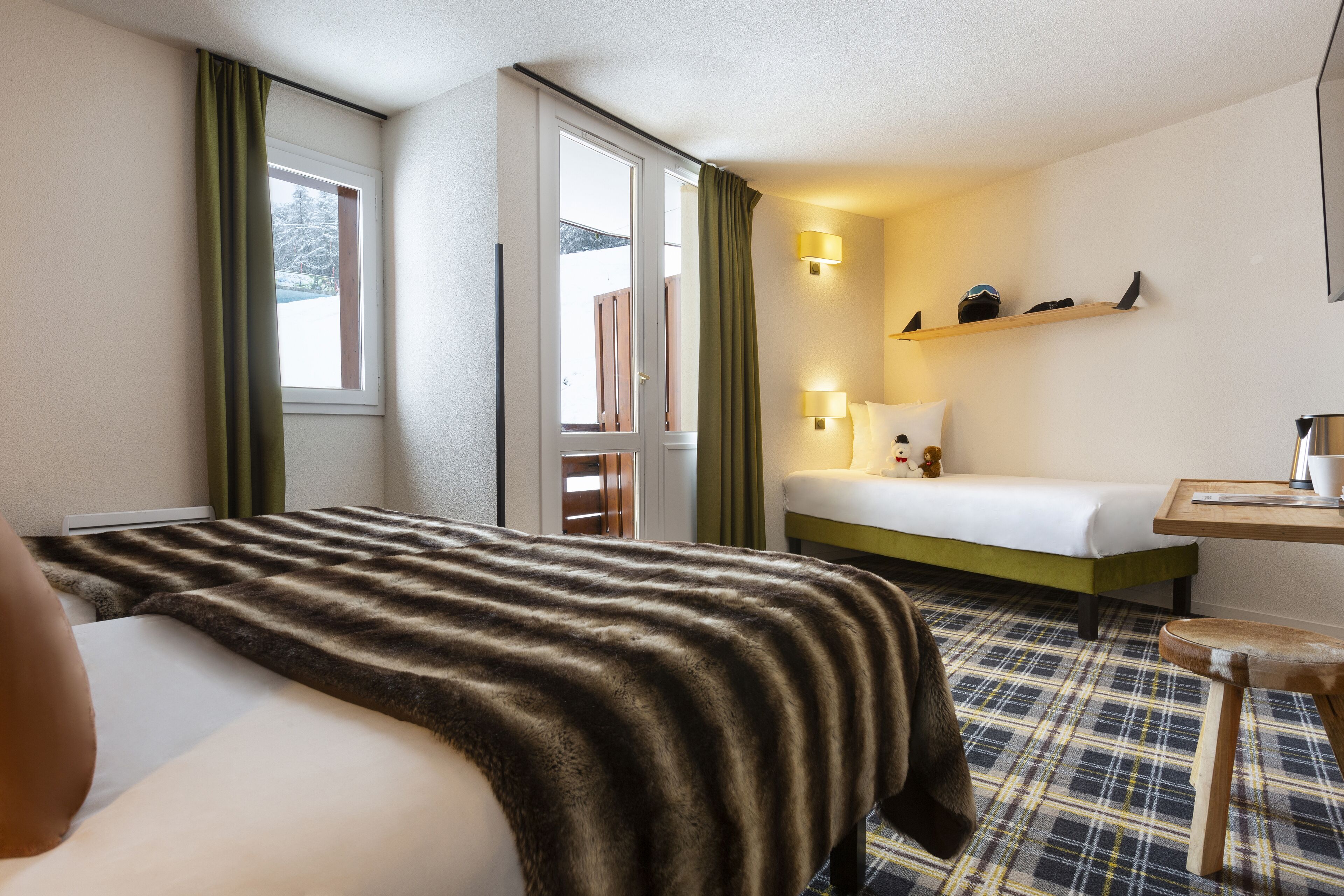 Une chambre d’hôtel avec un lit, un bureau, une chaise, une étagère avec quelques objets et une fenêtre avec des rideaux.