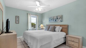 A bedroom with a bed, a nightstand, a dresser, a ceiling fan, a window with curtains, and a painting on the wall. - Gulf Front, Stunning Views, Great Location + Free Attraction Tickets! (Seacrest)