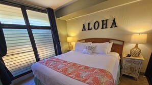 Et hotelværelse med en seng, et natbord med en lampe, et vindue med persienner og en v æg med ordet "ALOHA".