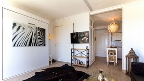 Una sala de estar con una obra de arte grande de estampado de cebra, un televisor, una mesa de centro de madera con un objeto decorativo, un tapete de cuero negro y una zona de cocina con un taburete y un farol.