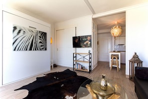A living room with a large zebra print artwork, a television, a wooden coffee table with a decorative item, a black leather rug, and a kitchen area with a stool and a lantern.