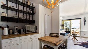 Una cocina moderna con armarios blancos, encimera de madera y una isla central. Hay un comedor con un sofá y vistas al exterior.