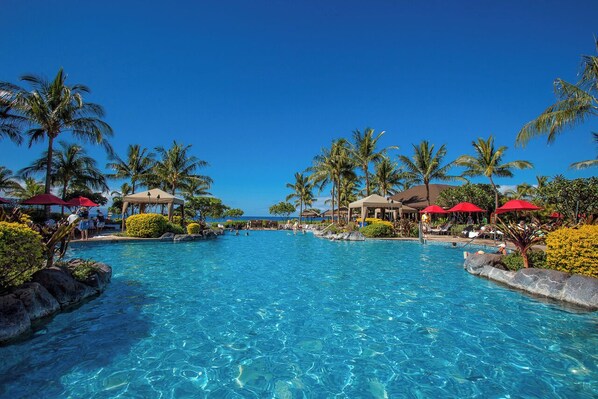 A clear blue pool surrounded by palm trees and red umbrellas, with lounge chairs and a view of the ocean.
