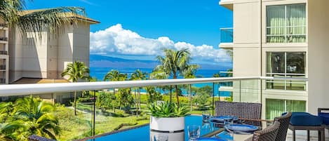 Un balcon avec une table en verre, dressée pour le dîner, donnant sur une piscine et une végétation luxuriante.