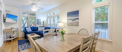 A dining area with a wooden table, chairs, a vase with flowers, a TV on a stand, a blue sofa, and a window with blinds.