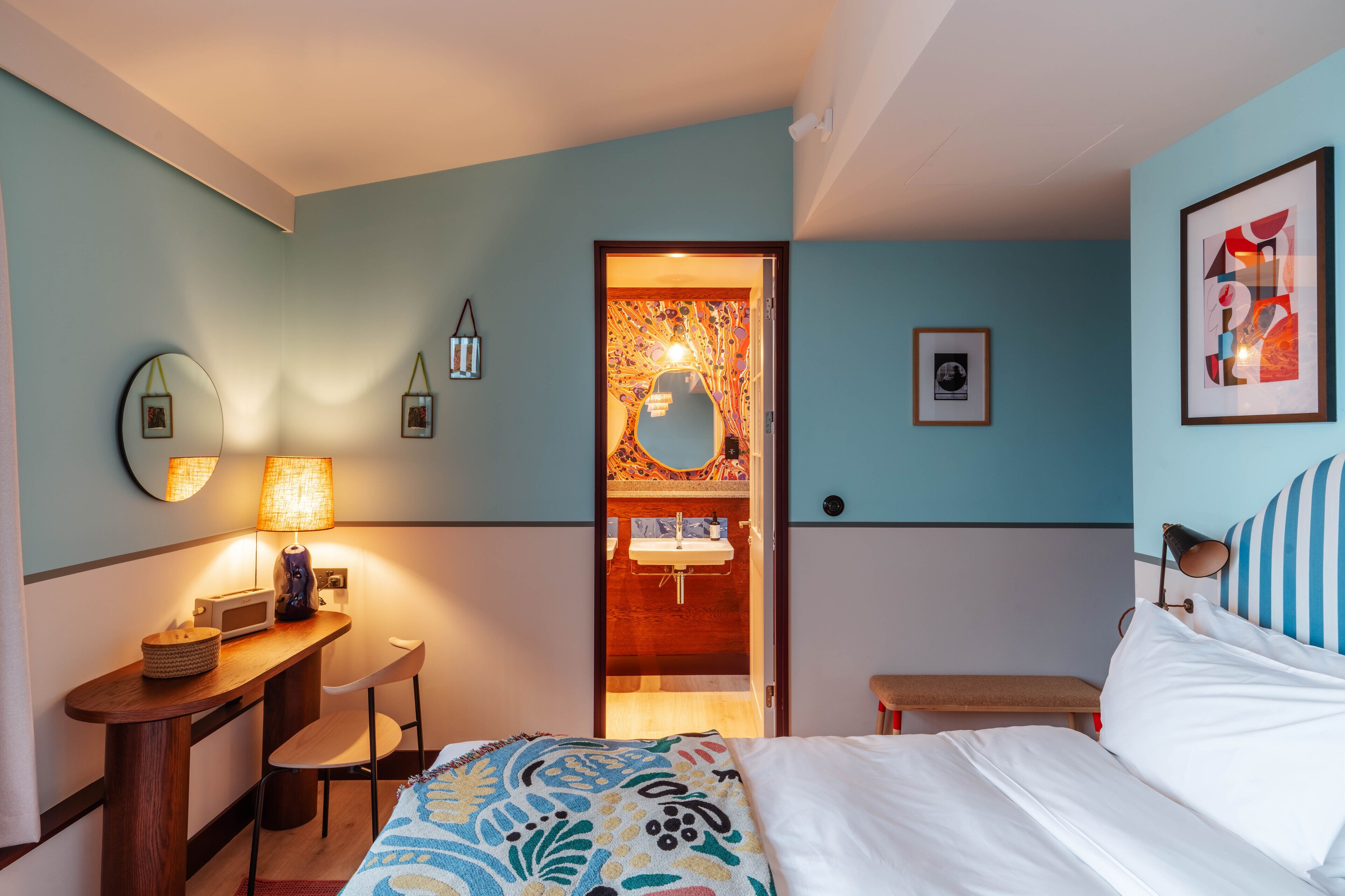 Ein Hotelzimmer mit Bett, Schreibtisch mit Lampe, Stuhl, kleinem Tisch und einem durch eine offene Tür einsehbaren Badezimmer.