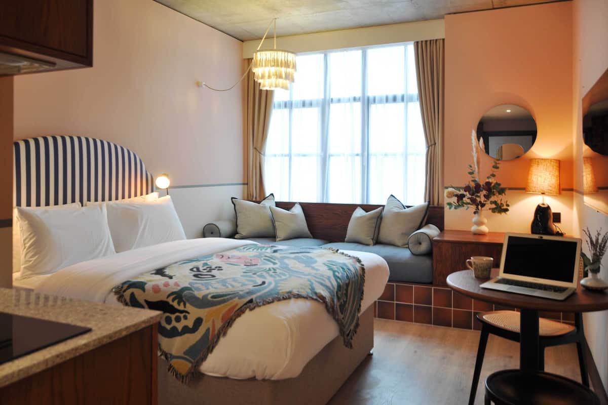 A hotel room with a bed, a desk with a laptop, a window with curtains, and a chandelier.