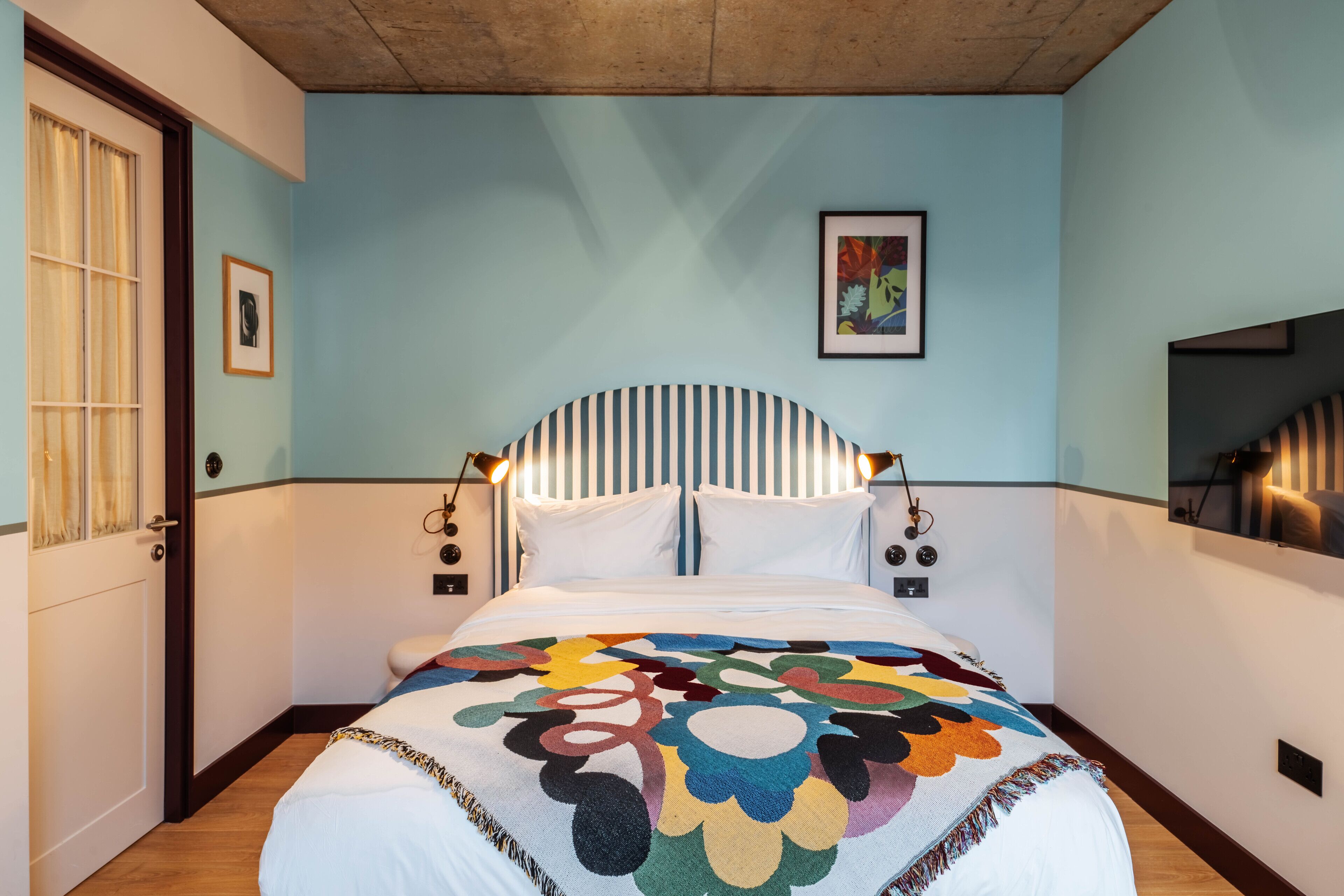 Une chambre à coucher avec un lit, une fenêtre avec des rideaux, un tableau encadré au mur et un luminaire au-dessus du lit.