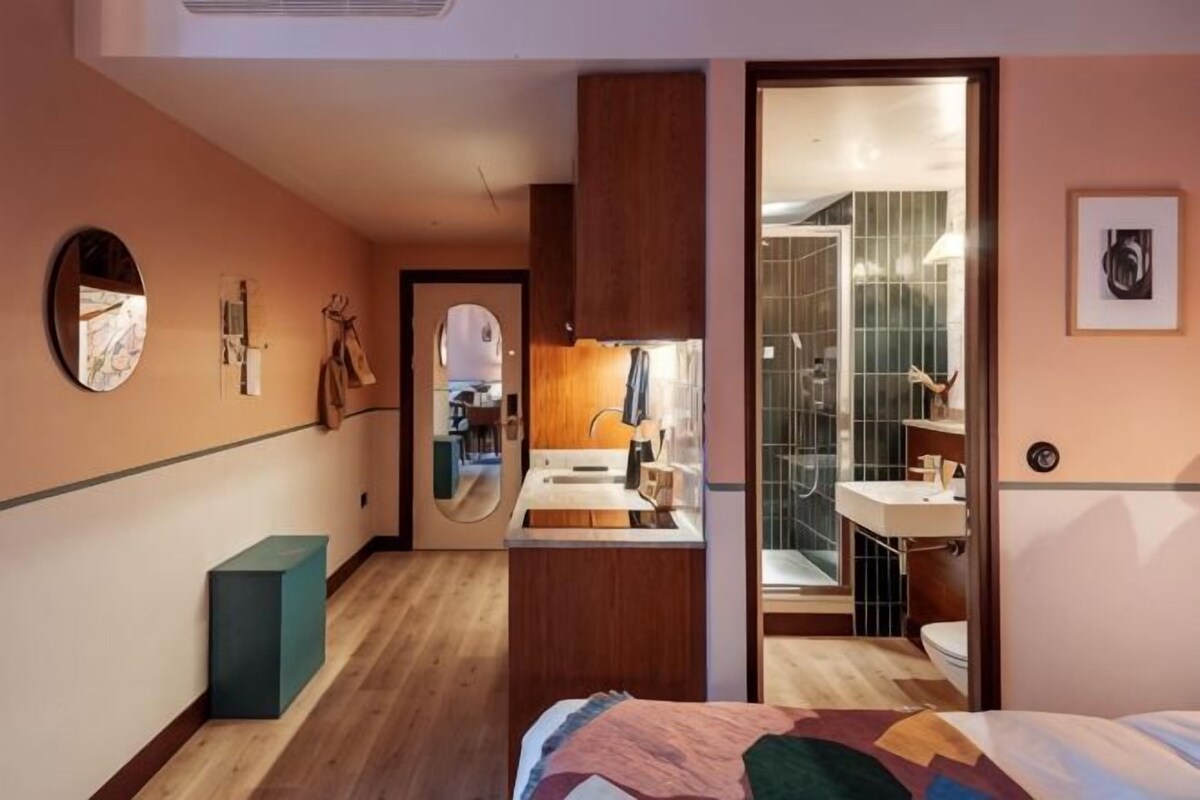 A bedroom with a bed, a bedside table, a mirror, and a door leading to a bathroom with a sink and mirror.