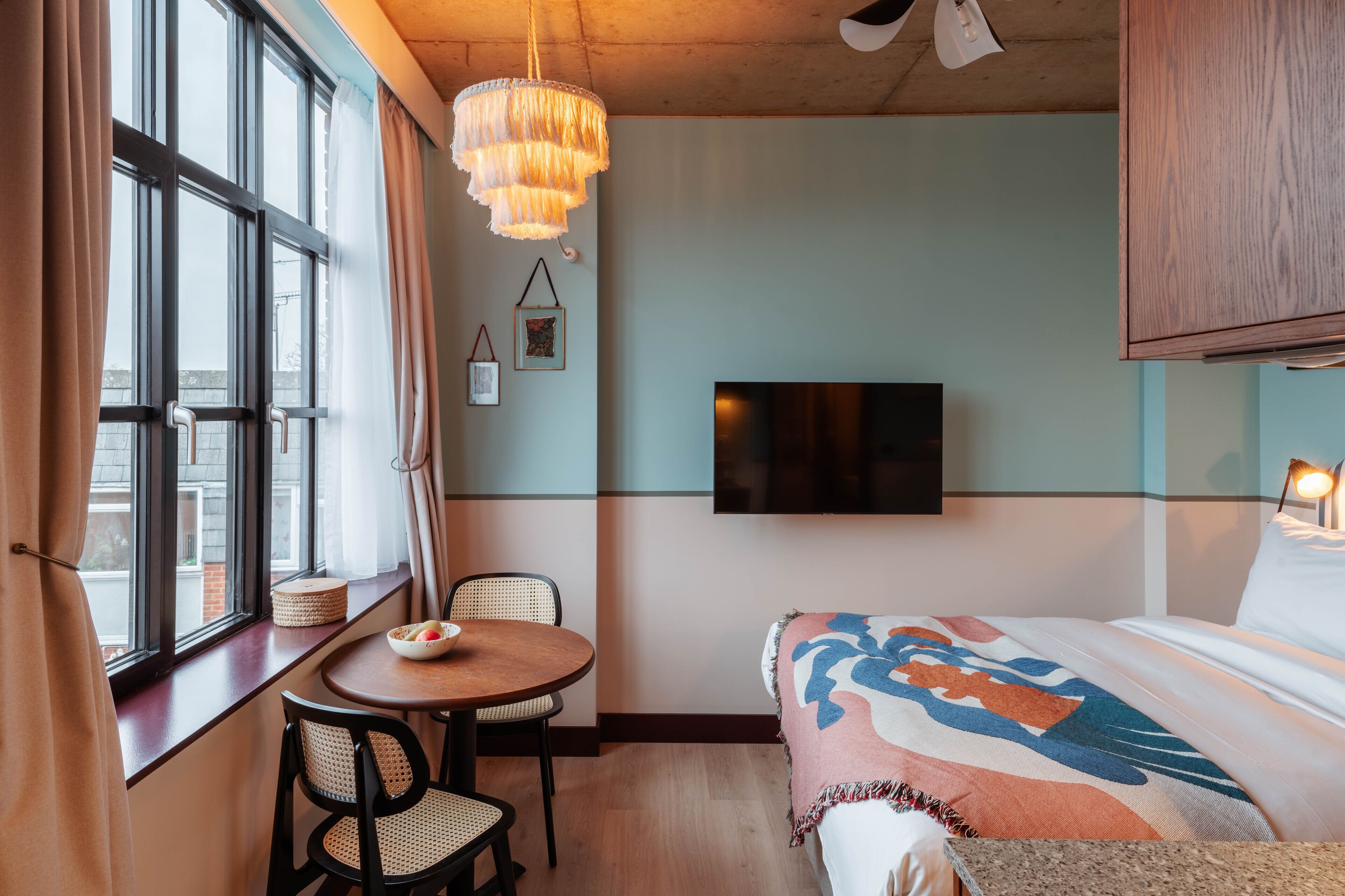 A hotel room with a bed, a TV mounted on the wall, a dining table with chairs, and a window with curtains.
