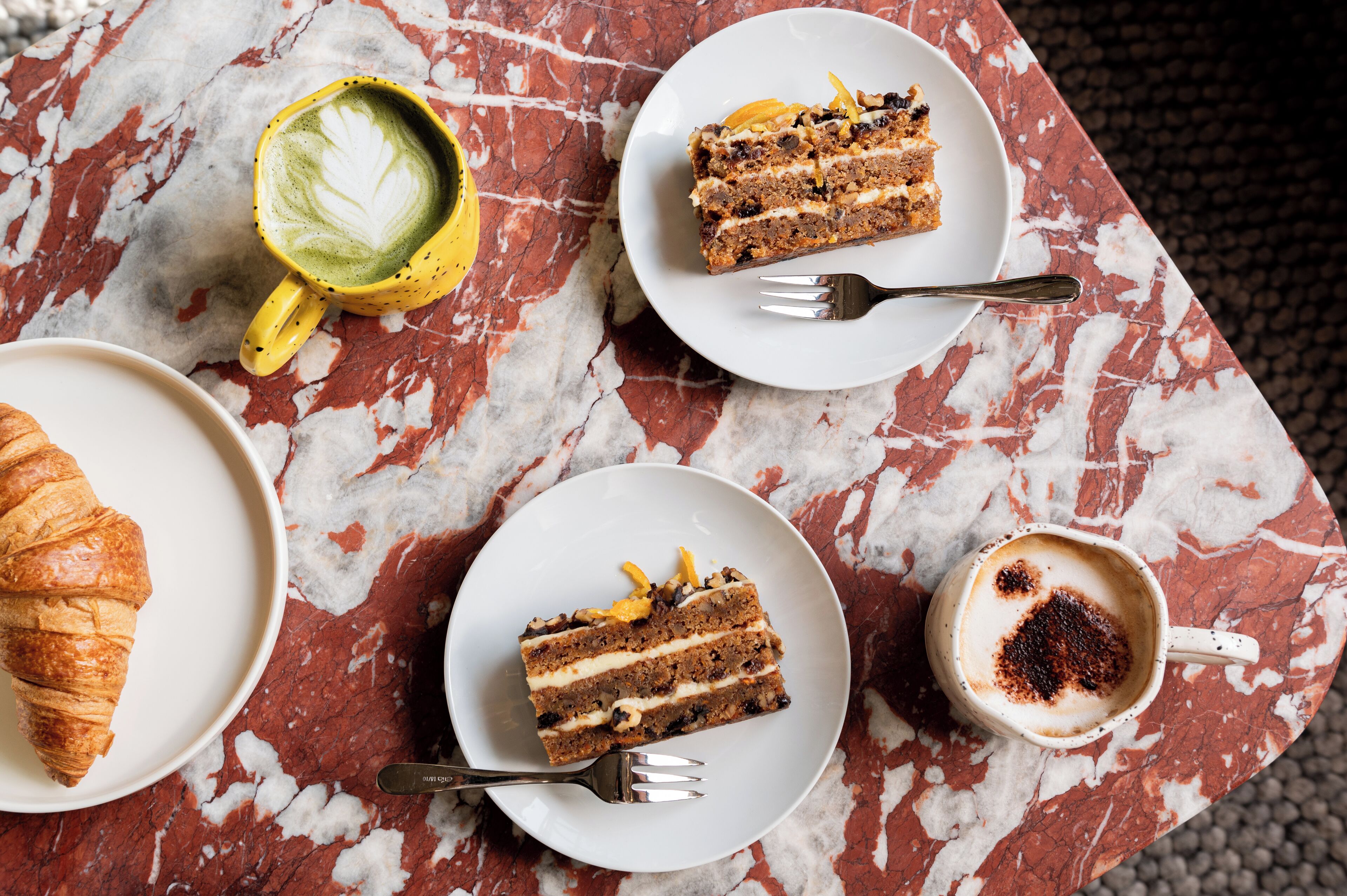 A table with a croissant, a slice of cake, a cup of coffee, and a latte with latte art.