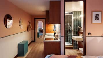 A hotel room with a wooden floor, a bed with a colorful blanket, a bathroom with a shower and sink, and a hallway leading to a dining area.