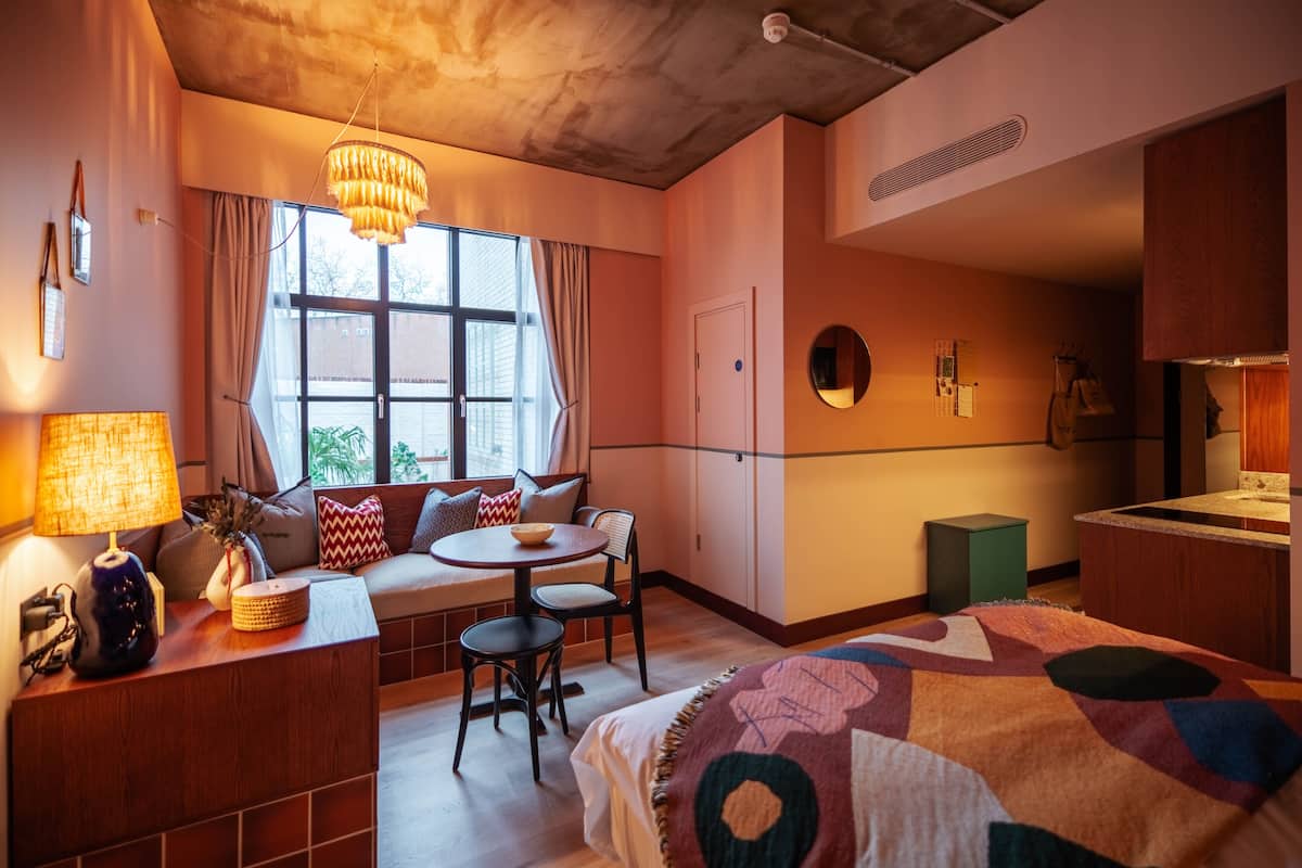 A cozy hotel room with a large bed, a small round table, a sofa with cushions, a window with curtains, and a chandelier.