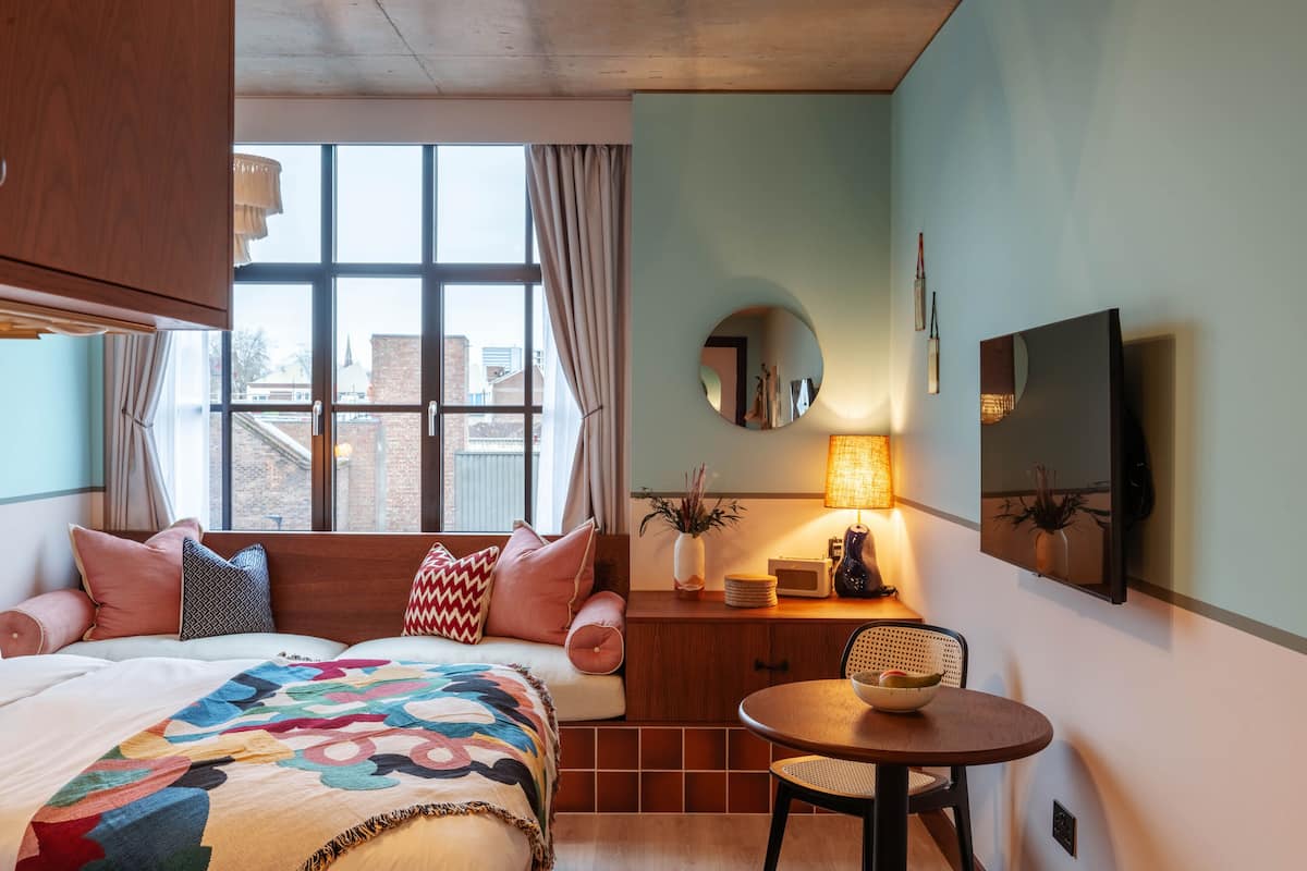A bedroom with a large bed, a window with curtains, a bedside table with a lamp, a round mirror, and a television on the wall.