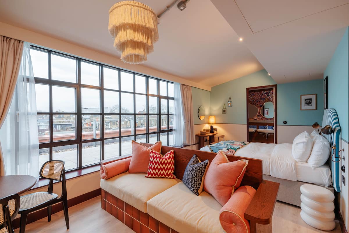 A modern hotel room with a large window, a sofa, a bed, and a dining area.