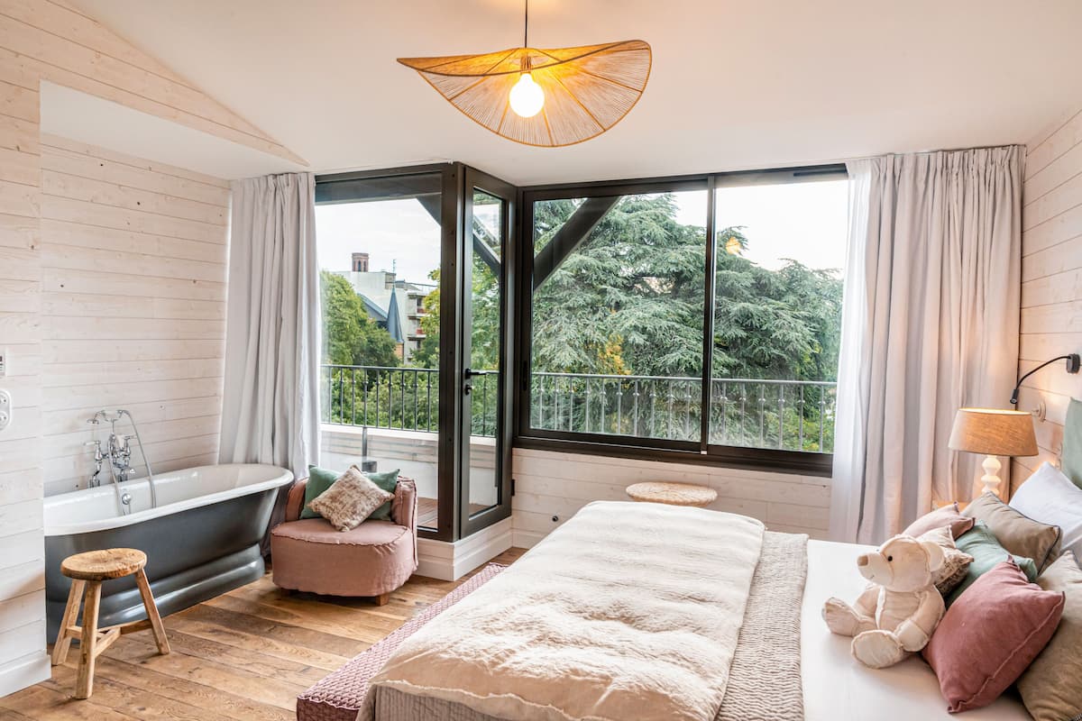 A bedroom with a large window, a bathtub, a bed, and a bedside lamp.