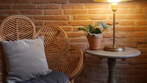 A room with a brick wall, a round wooden table with a lamp, a potted plant, and two wicker chairs with cushions. - Hôtel SOCLO (Toulouse)