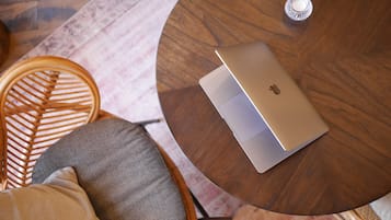 A round wooden table with a laptop, a woven chair, and a cushion.