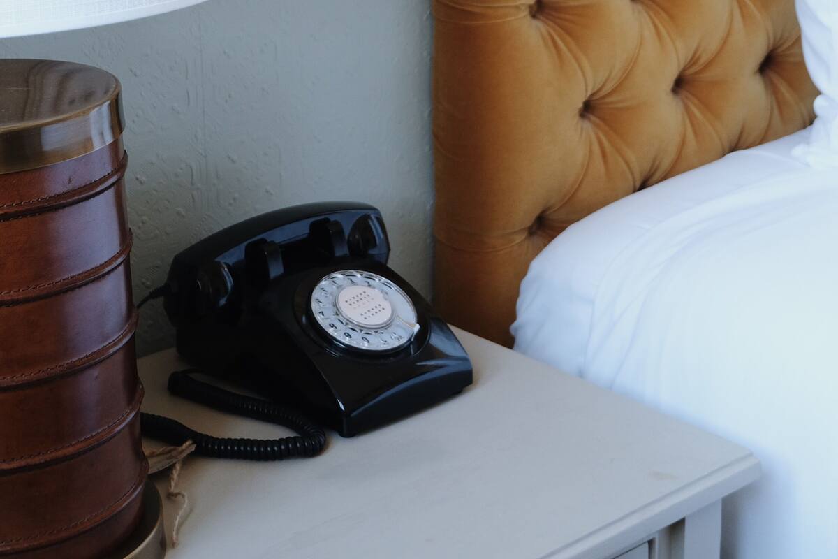 A bed with a tufted headboard, a white pillow, a black rotary phone, and a white lamp on a bedside table.