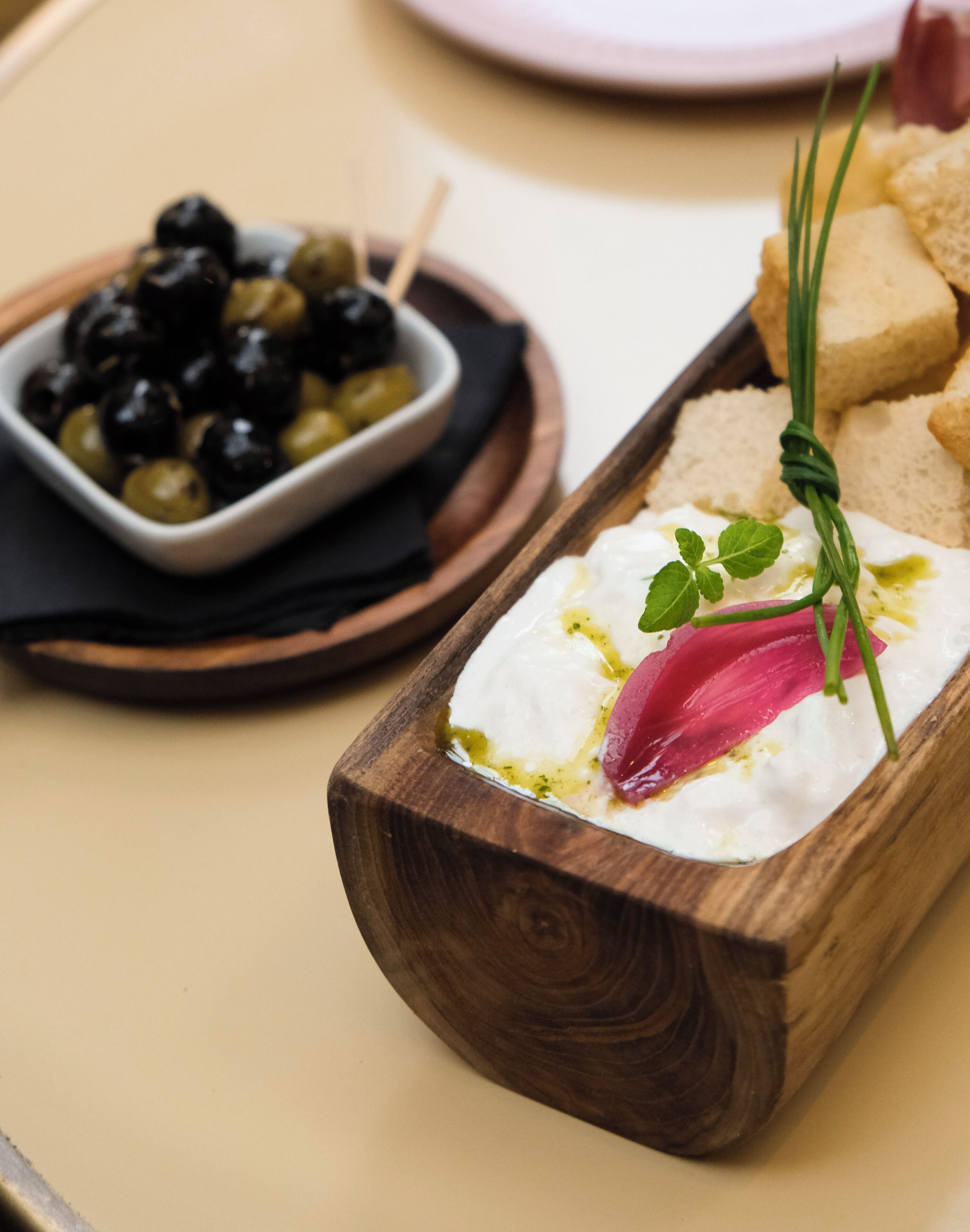 A wooden serving tray with a white creamy spread, garnished with a red vegetable and green herbs, accompanied by a bowl of olives and bread on a table.