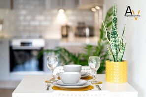Une table à manger dressée avec des bols blancs, des assiettes dorées et des verres à vin, et un vase jaune contenant une plante.
