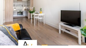 A modern living room with a flat-screen TV, a dining area with a white table and chairs, and a kitchen in the background.