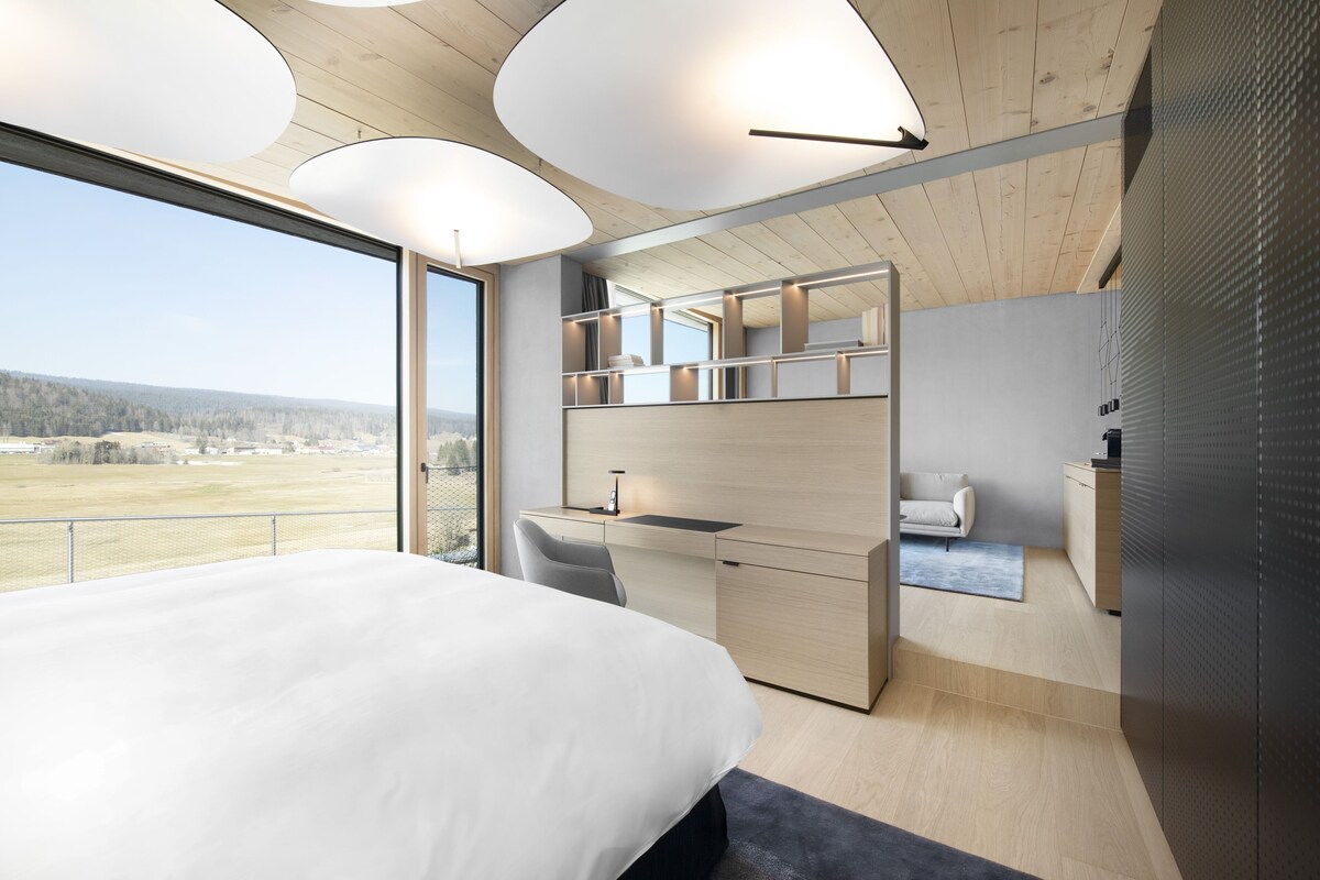A modern bedroom with a large bed, a desk, a chair, and a view of the outdoors.