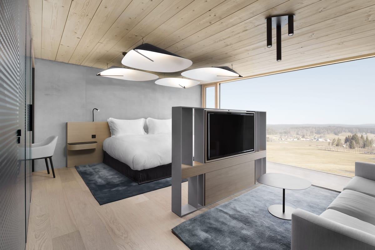 A modern bedroom with a large bed, a TV, a sofa, and a view of the countryside.