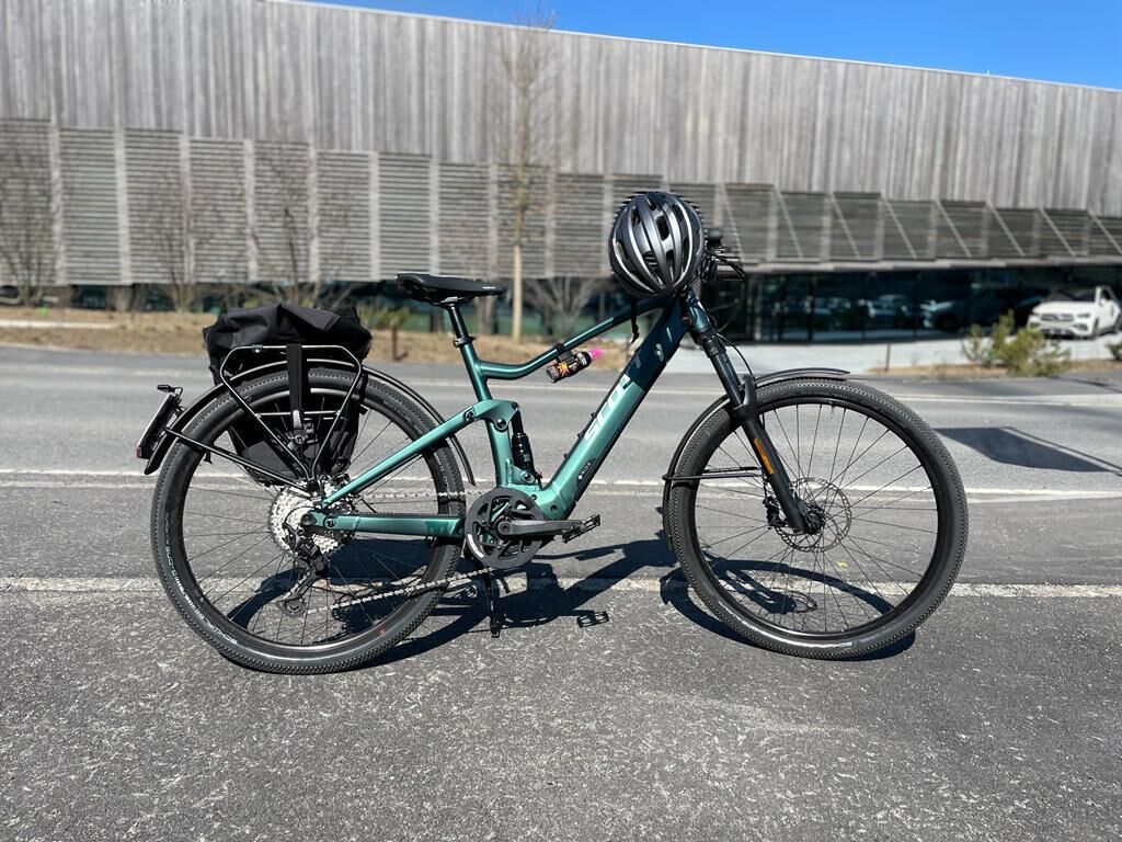 A teal bicycle with a black seat and front rack, parked on a paved surface with a modern building in the background.