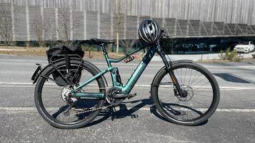 A teal bicycle with a black seat and front rack, parked on a paved surface with a modern building in the background.