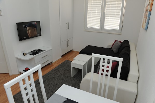 Un salon moderne et bien éclairé, avec un téléviseur à écran plat fixé au mur, un canapé noir agrémenté de coussins rouges et gris, une table à manger blanche et une chaise blanche.