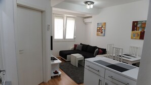 Ett kompakt bostadsutrymme med en soffa, ett litet bord, en liten köksdel och ett fönster med persienner.