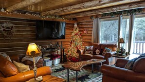 A cozy cabin interior with a wooden ceiling, leather sofas, a television, a decorated Christmas tree, and a large window with a view of trees.
