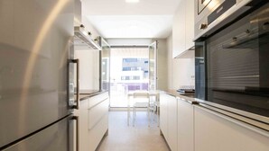 A narrow kitchen with white cabinets, a built-in oven, and a window overlooking a balcony.