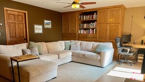 Una sala de estar con un sofá modular, una estantería de madera, un escritorio con una computadora y un ventilador de techo.