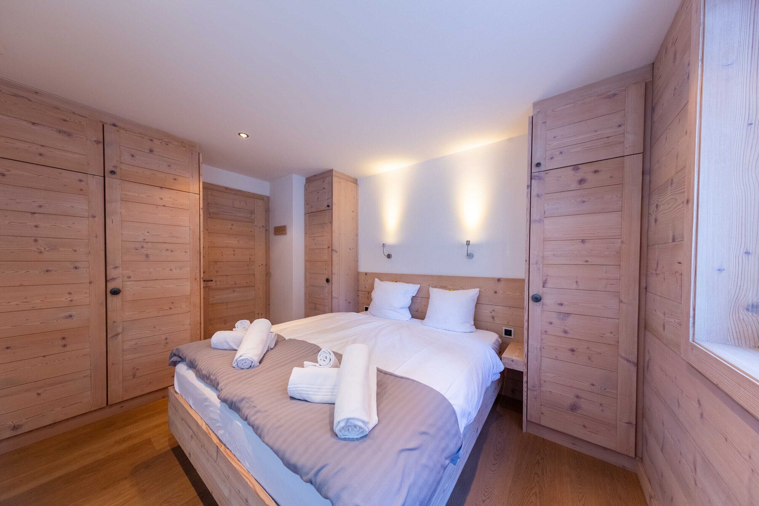 A bedroom with a large bed, wooden wardrobe, and wall-mounted lighting.