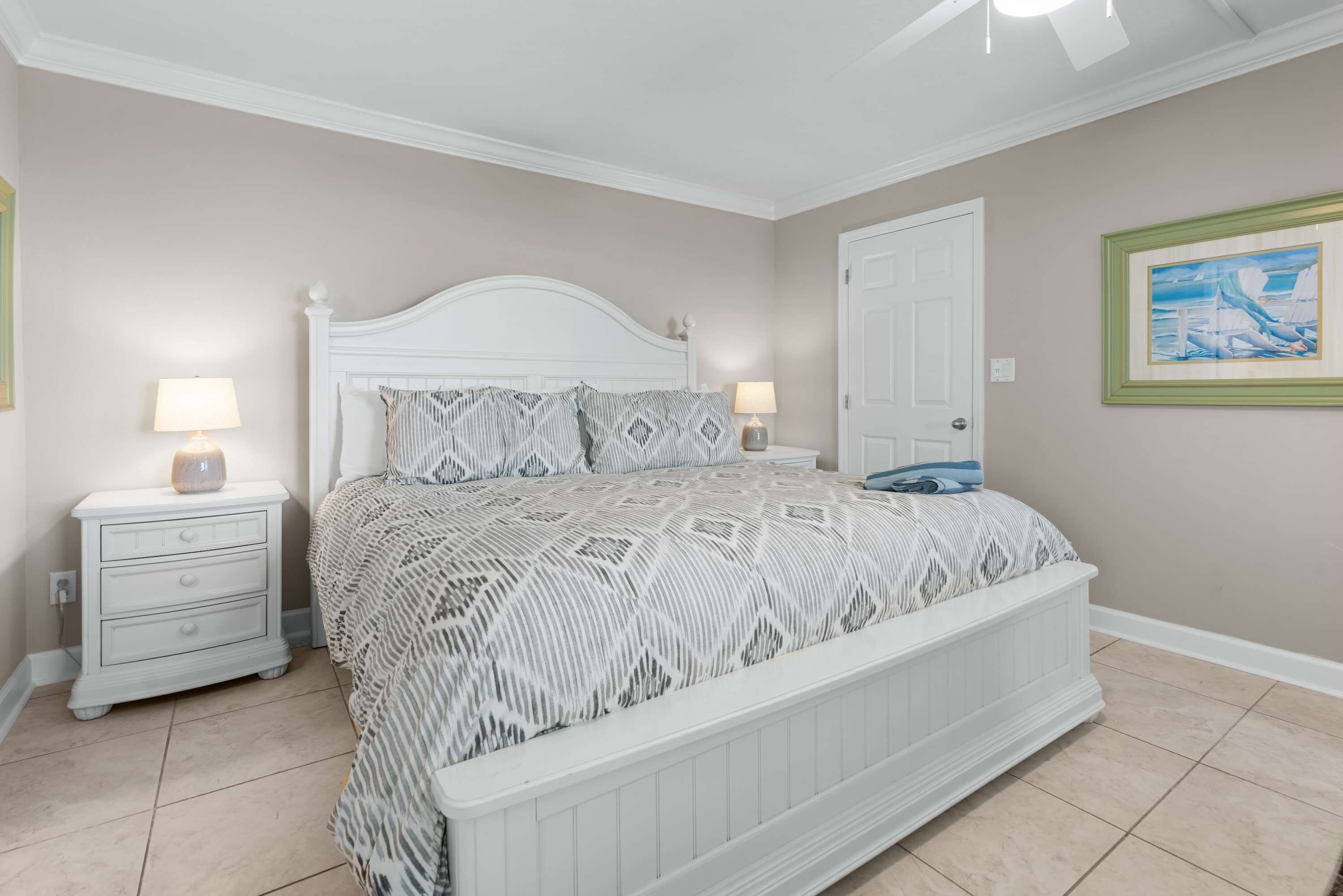 A bedroom with a white bed, two bedside tables with lamps, a door, and a framed picture on the wall.