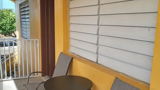 A balcony with a table and two chairs, a window with blinds, and a view of a parked car and greenery.