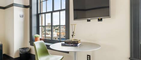 A room with a large window, a round table with books, a green chair, a potted plant, and a mounted TV.