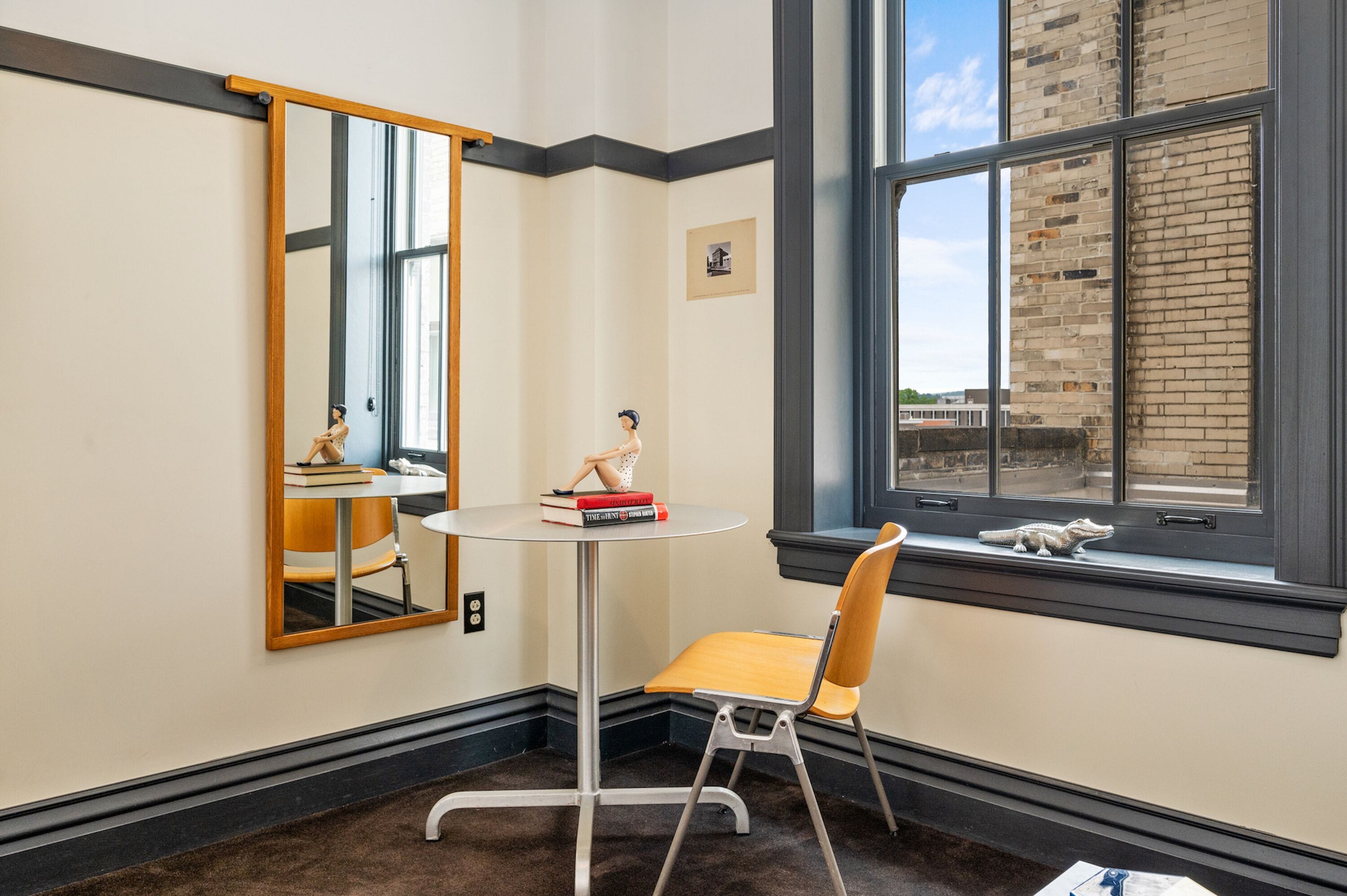 Una habitación pequeña con una mesa redonda, una silla, un espejo grande y una ventana que da a un edificio de ladrillo.