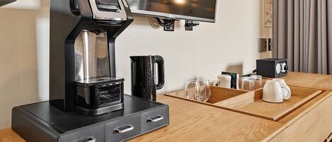 A coffee machine on a wooden counter with a TV above it, a coffee pot, and a glass of water.