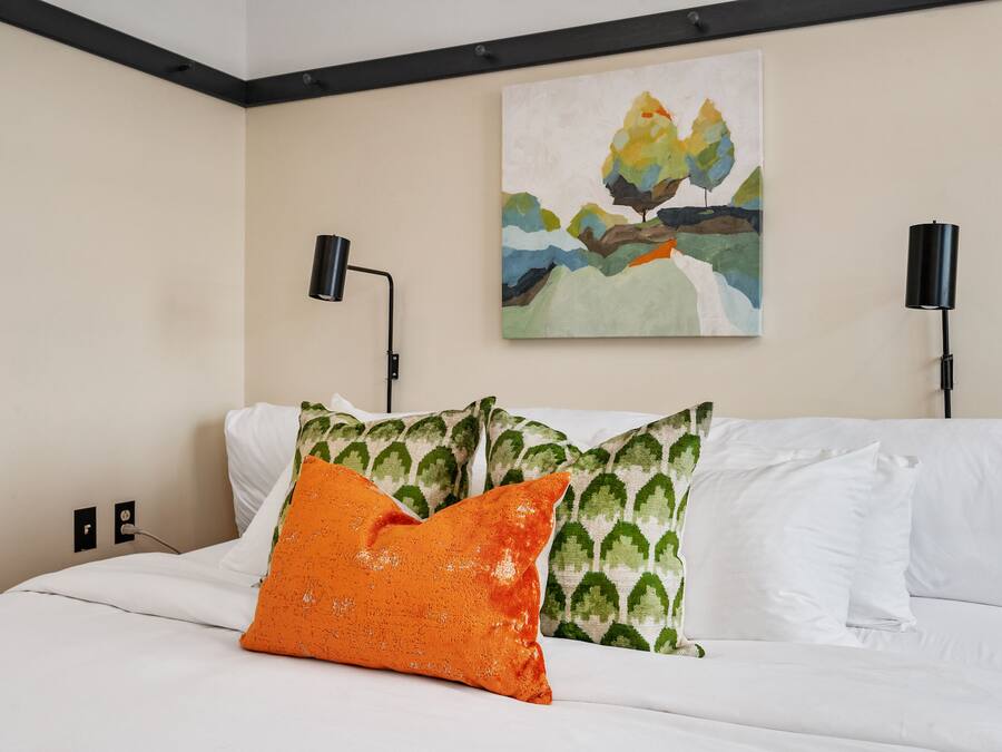 A neatly made bed with white bedding and decorative pillows, a painting on the wall, and two wall-mounted lamps.