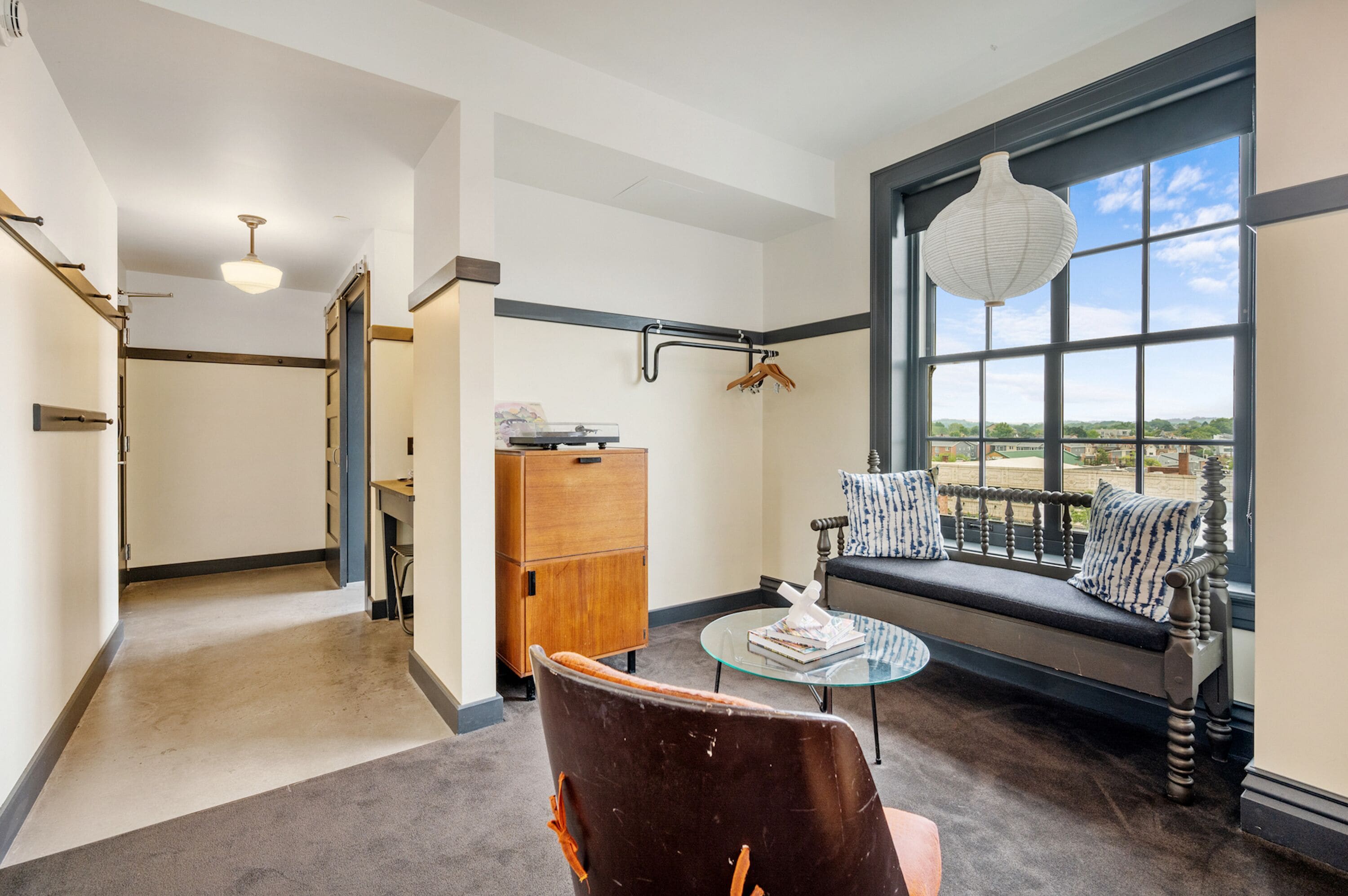 A room with a large window, a wooden cabinet, a bench with cushions, and a glass table.