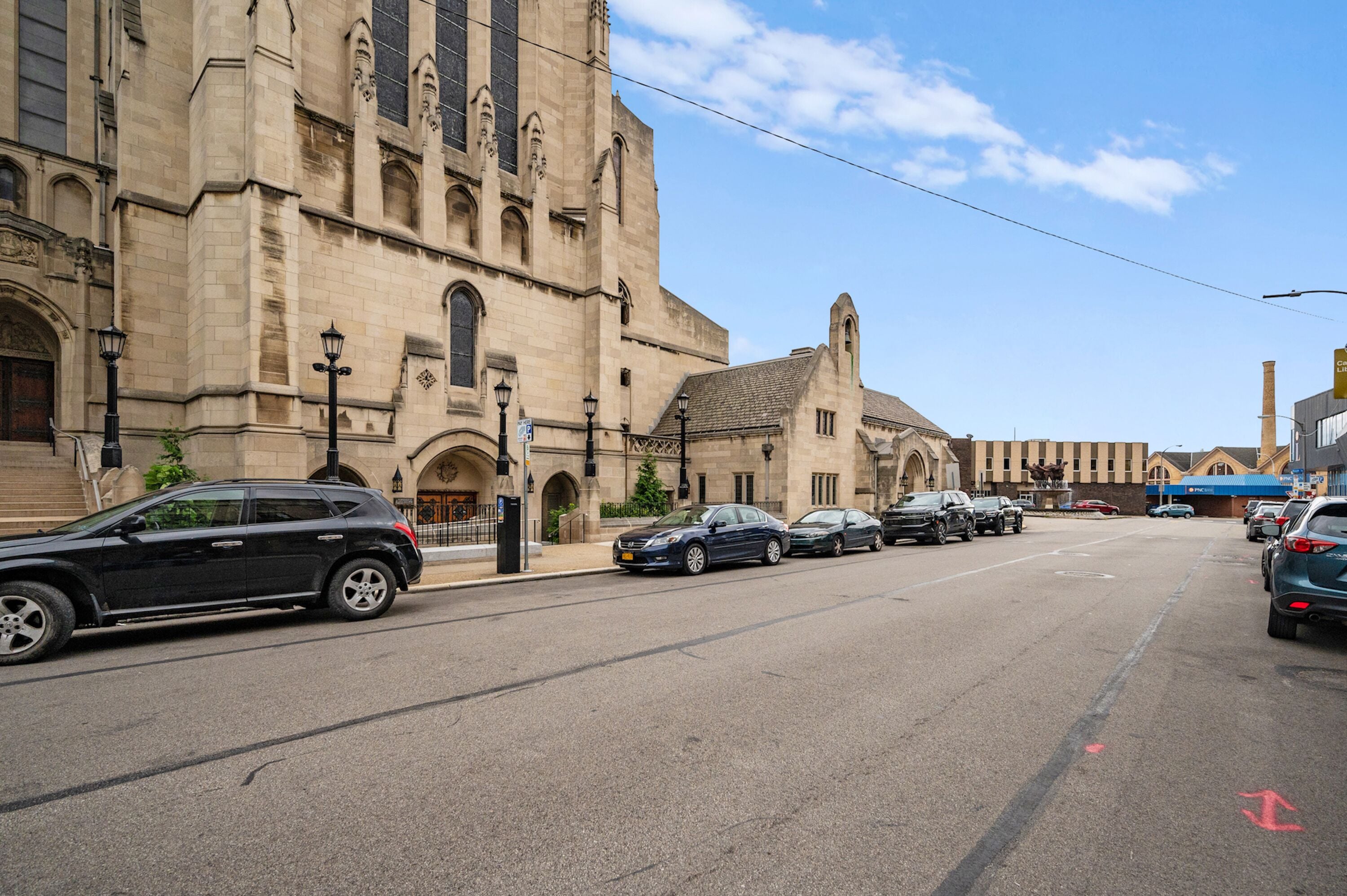 A street with parked cars and a large historic building with arched windows and a clock tower.