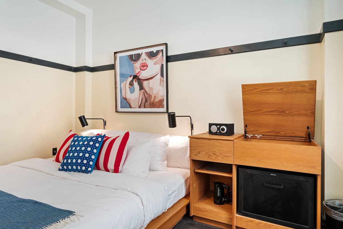 A hotel room with a bed, a wooden nightstand with a radio, a coffee maker, and a framed picture on the wall.