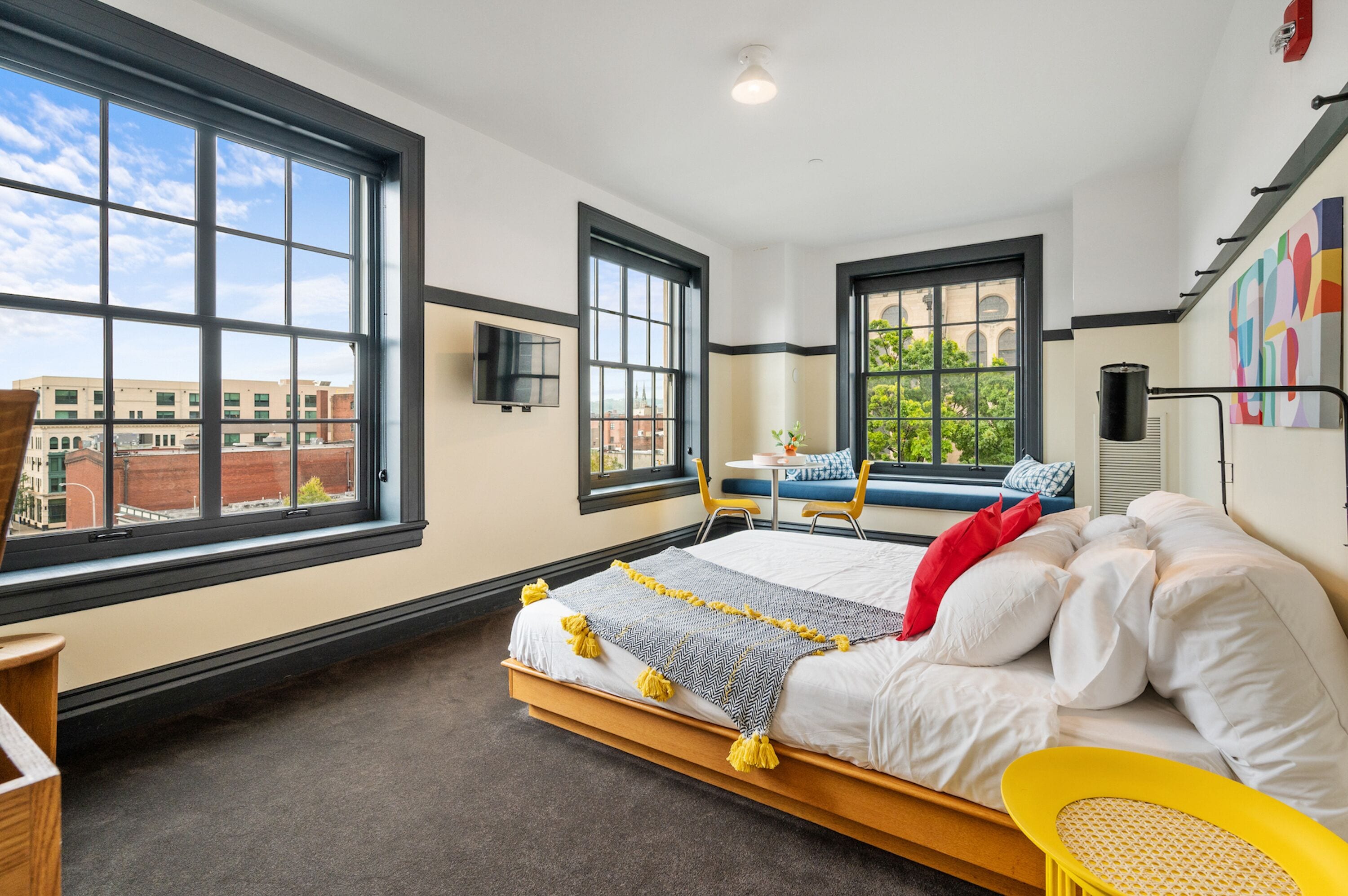 A modern hotel room with a large bed, a desk, and a view of the cityscape through large windows.